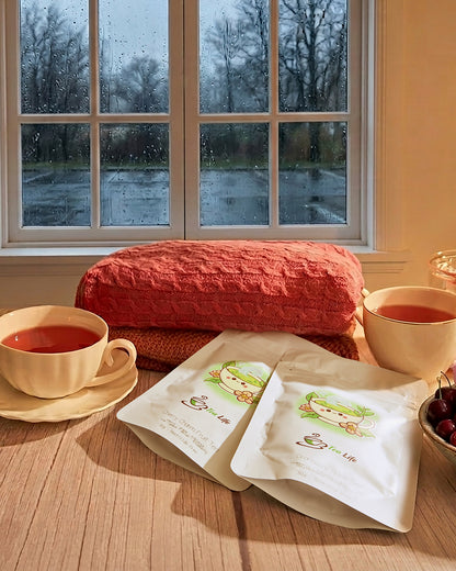 Two white pouches of herbal tea with cartoon graphics are displayed on a wooden table in a cozy room setting, with two cups of tea, a red knit pillow, and a window showing a rainy day outside.
