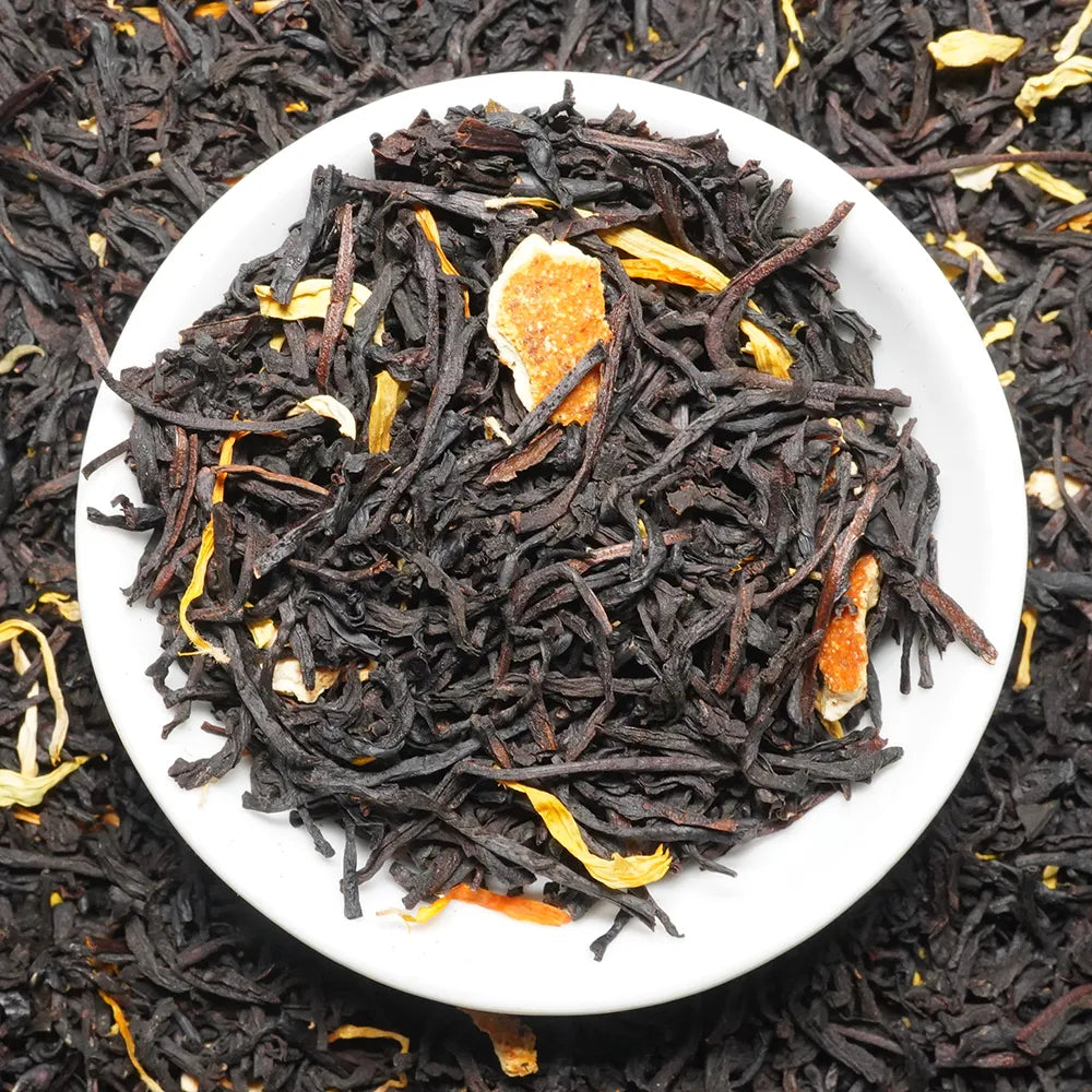 Dark loose-leaf tea blend in a white round dish, with visible dried citrus peel pieces and small yellow botanical accents, photographed from above against a matching tea-filled background.

